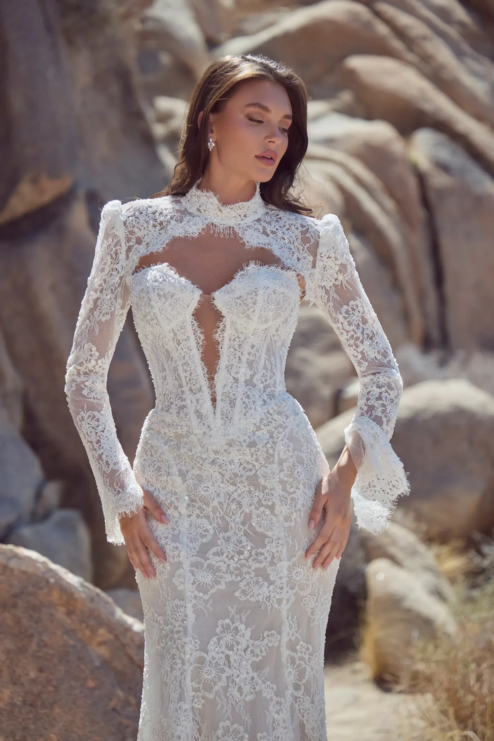 A woman in an elegant, long-sleeved lace wedding dress stands outdoors against a rocky backdrop. She exudes a serene and graceful demeanor.
