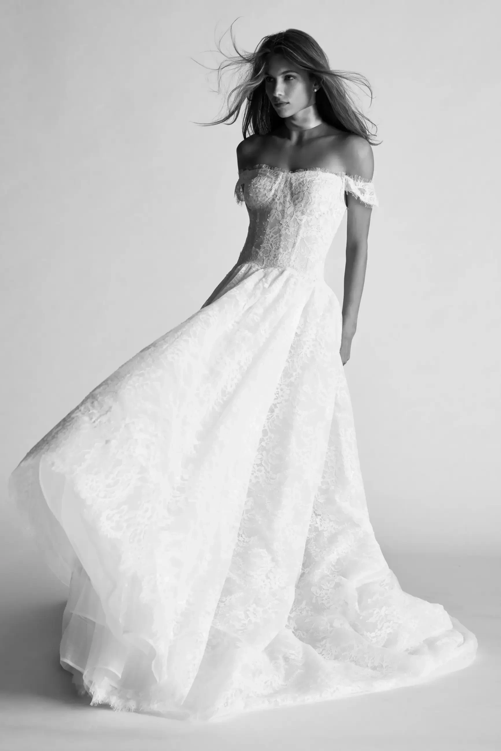 Elegant bride in off-the-shoulder lace gown with flowing skirt, hair gently windswept. Monochrome setting conveys timeless, graceful allure.