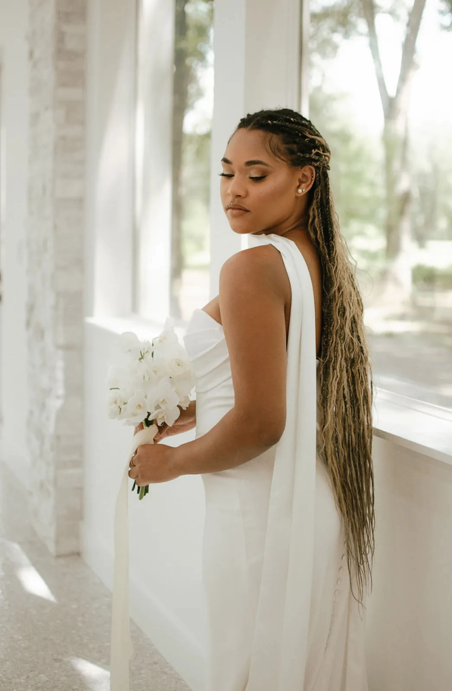 Bride in an elegant white gown holds a bouquet of white flowers, standing by a bright window. Her long, braided hair adds graceful detail.