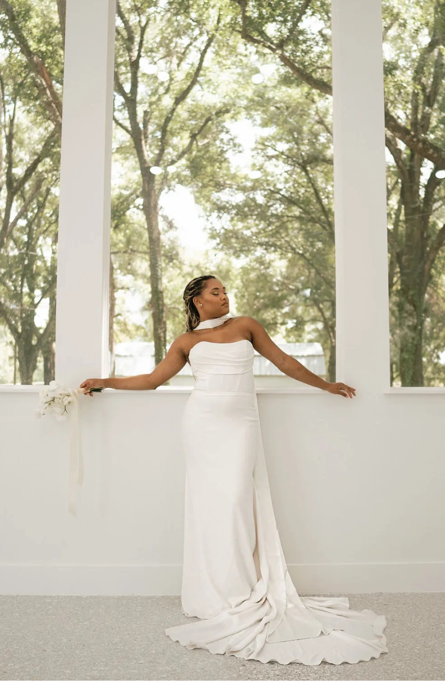 A woman in a strapless white wedding dress stands gracefully in front of large windows with trees visible outside. The scene is serene and elegant.