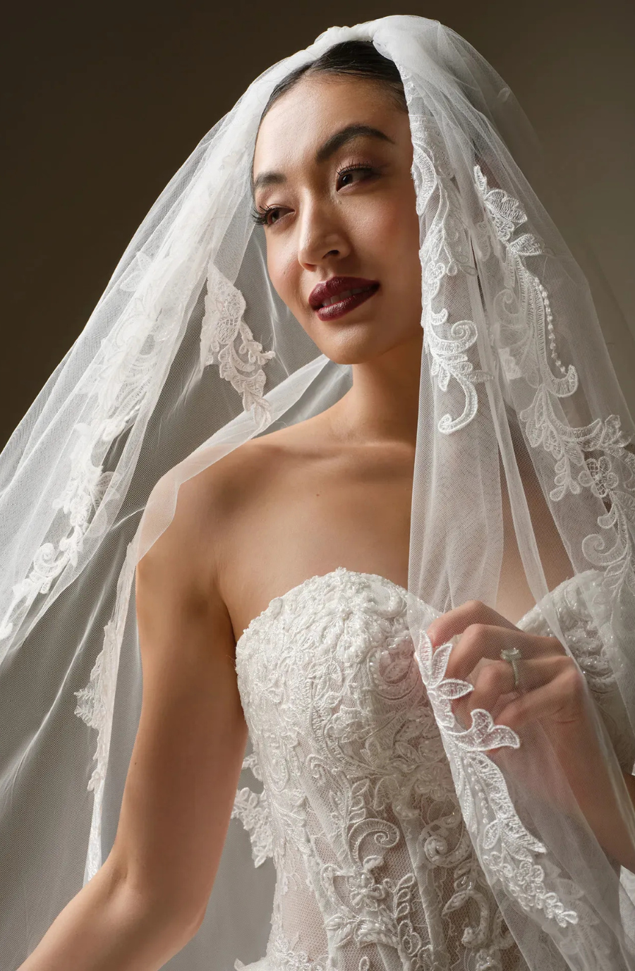 Bride in an elegant lace wedding dress and veil, gazing softly to the side. The intricate embroidery and serene expression convey romance and elegance.