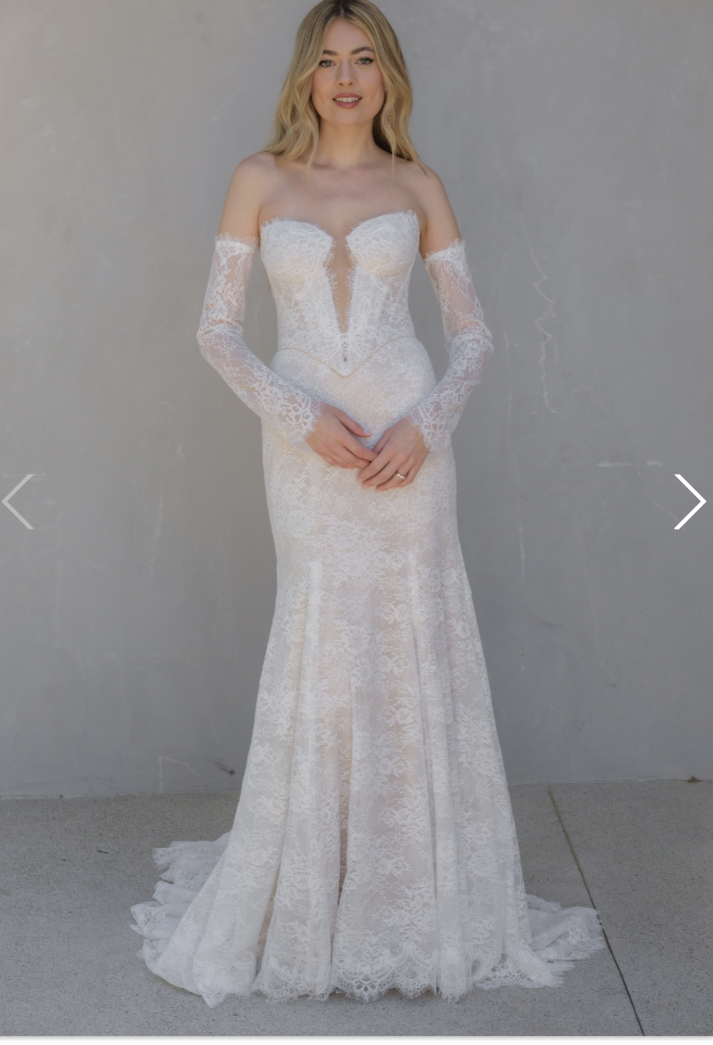 A model wearing a stylish off-the-shoulder lace wedding gown with long sleeves and a plunging neckline, standing against a neutral background.