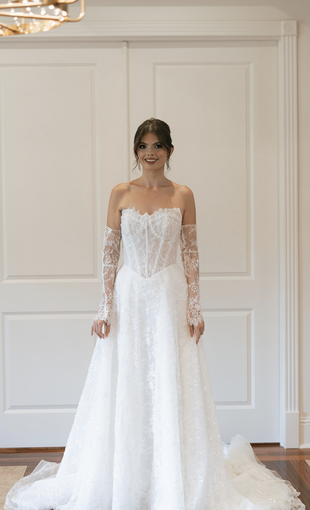 A bride wearing a strapless white wedding dress with lace gloves, standing in front of a white backdrop.