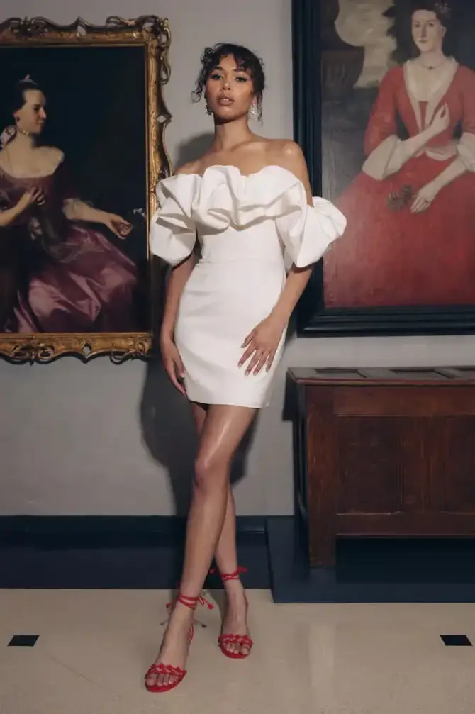 A woman in a white off-the-shoulder dress with ruffled sleeves stands elegantly in front of two paintings. She has long hair and is wearing red strappy heels, posing confidently with one leg slightly forward.