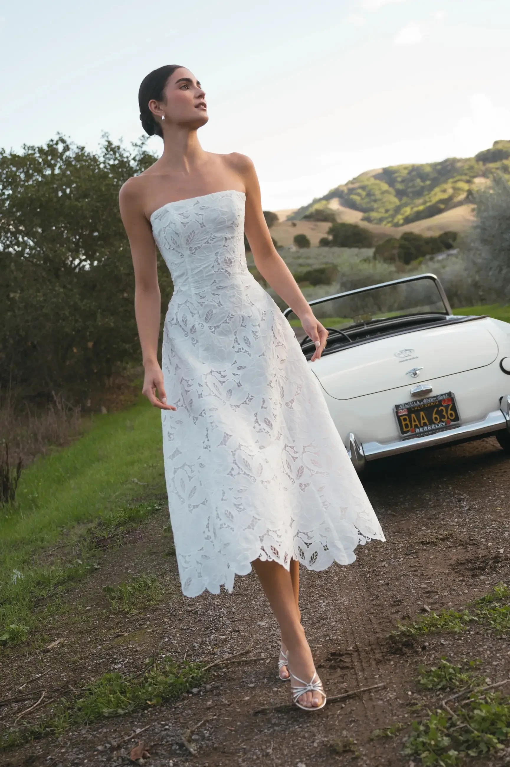 A woman in a strapless white dress stands on a dirt path, holding a small black purse, with green hills and trees in the background. A vintage car is parked nearby.