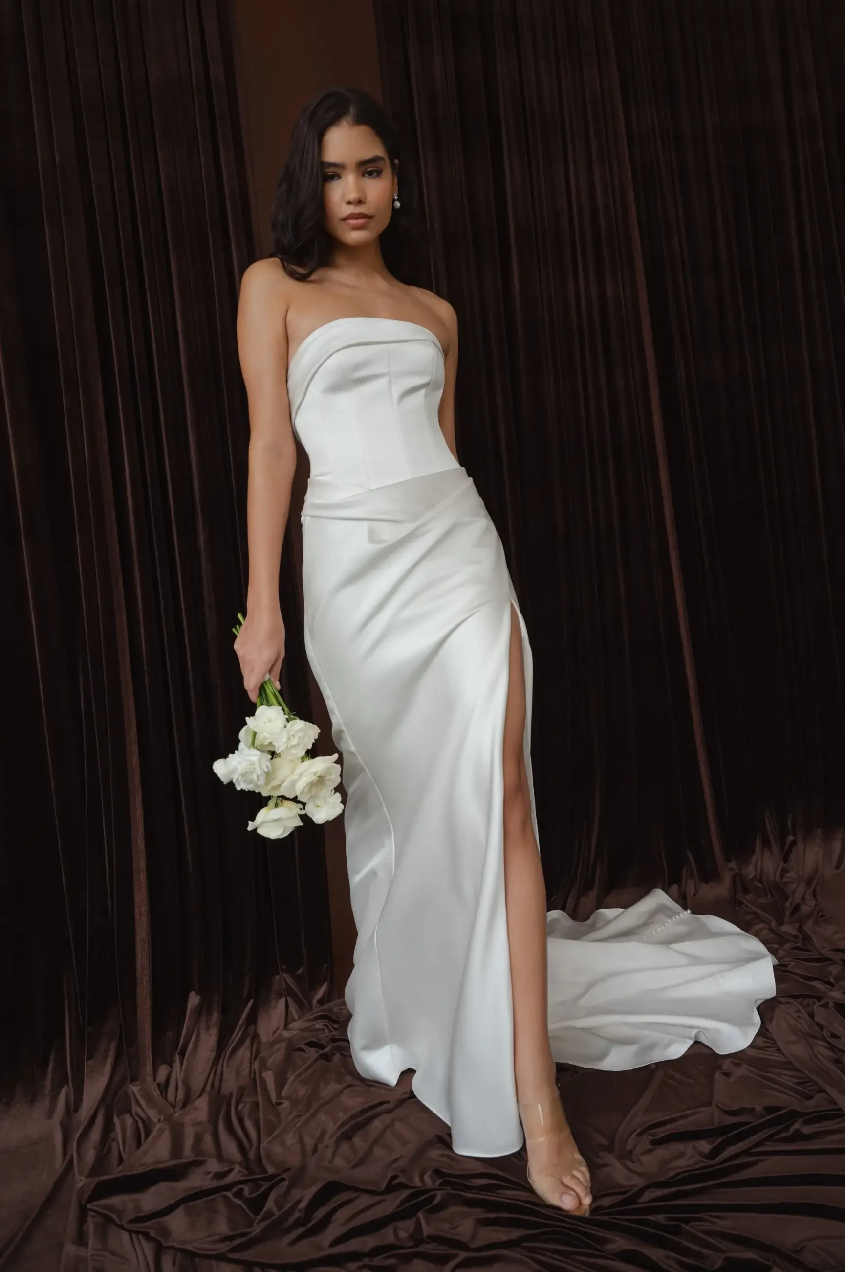Woman in a sleek, strapless white dress with a high slit stands on dark, flowing fabric. She holds a bouquet of white flowers, exuding elegance and sophistication.