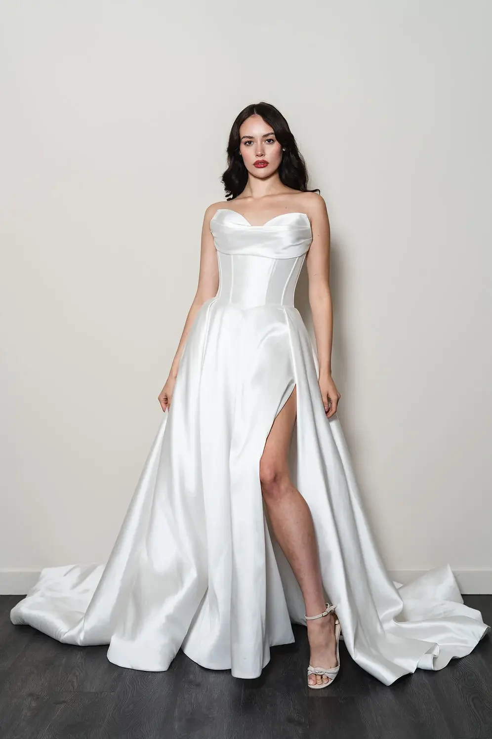 Woman in a white, strapless satin gown with a fitted bodice and thigh-high slit, standing confidently against a plain background. Tone is elegant and poised.