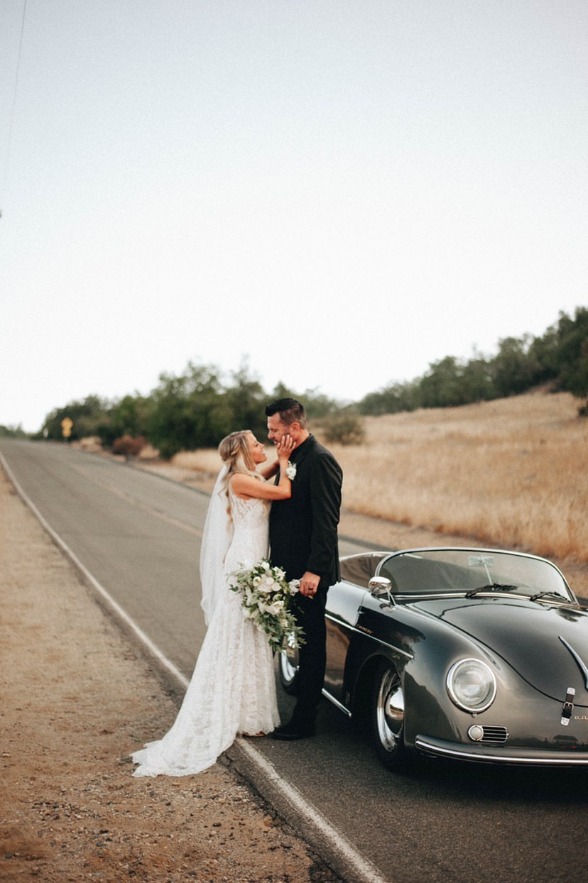 Сouple wearing a white gown and a black suit near the car